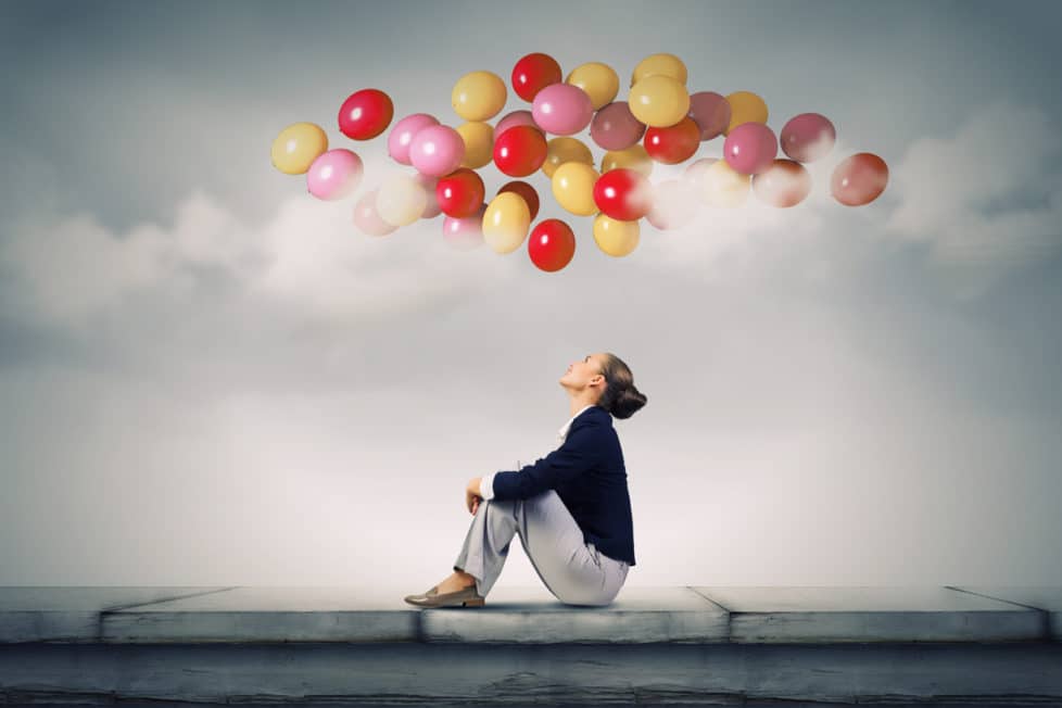 Frau sitzt auf Mauer und guckt zu bunten Luftballons über sich im Himmel