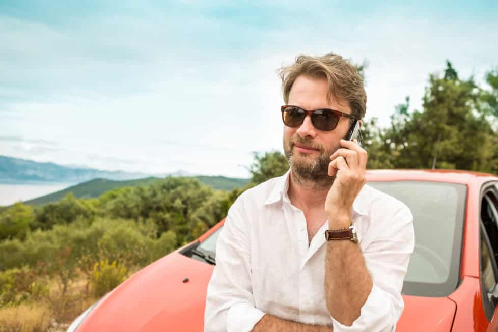 Mann telefoniert an Auto gelehnt im Urlaub mit Smartphone