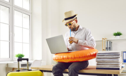 Business man in beach ring and summer hat sits on table in office with holiday suitcase, uses laptop, books online tickets to his annual leave vacation destination, and sips orange juice through straw