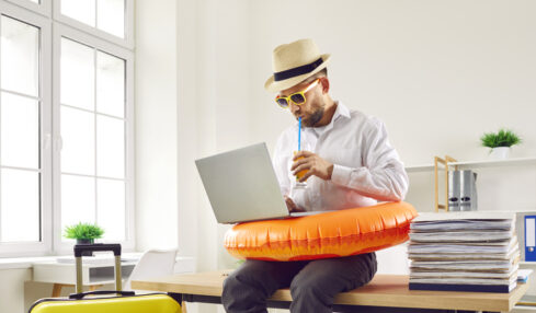 Business man in beach ring and summer hat sits on table in office with holiday suitcase, uses laptop, books online tickets to his annual leave vacation destination, and sips orange juice through straw